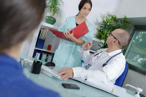 Nurse holding folder stood behind doctor in consultation with patient Stock Photos