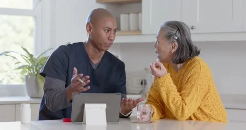 Nurse in scrubs pointing stylus explaining dose on tablet in kitchen as senior Vídeo Stock 345509450