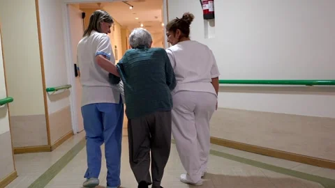 Nurses helping elderly woman walk in nursing home corridor Stock Footage 307592997