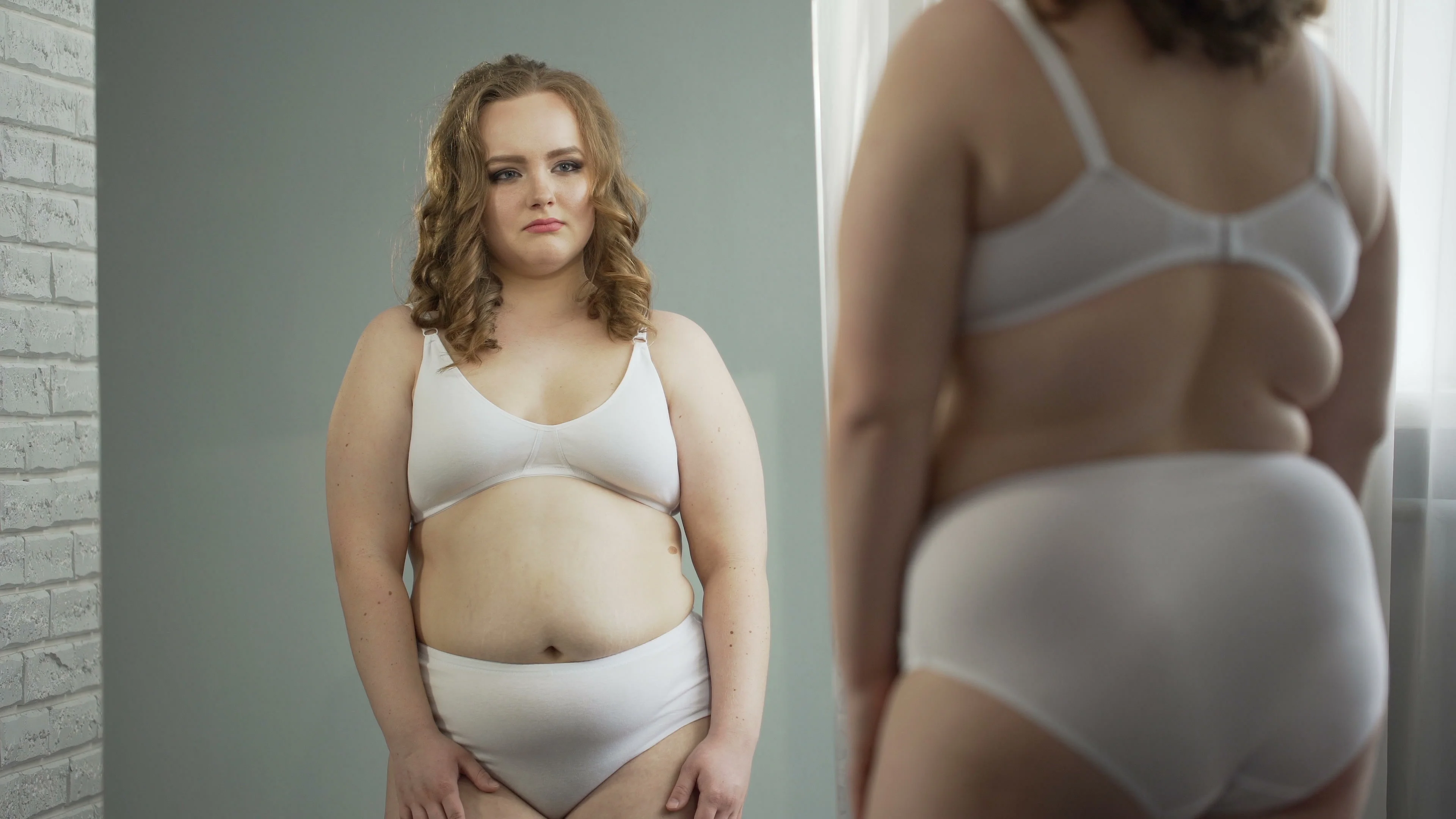 Obese lady in underwear looking at reflection in mirror ashamed of her fat body