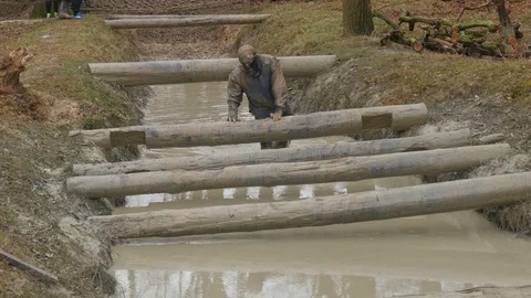 An obstacle course racer climbs over logs while in a muddy stream 動画素材 112005445