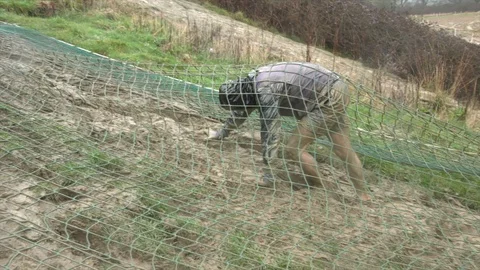 An obstacle course racer crawls under a cargo net up a hill Stock Footage 112003920