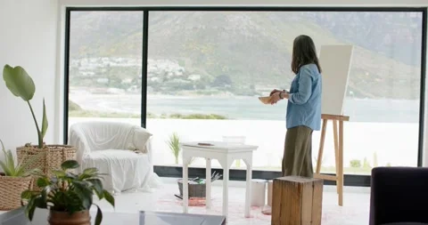 Ocean view inspiring painter adjusting palette and inspecting canvas by window Vídeos de archivo 342392420