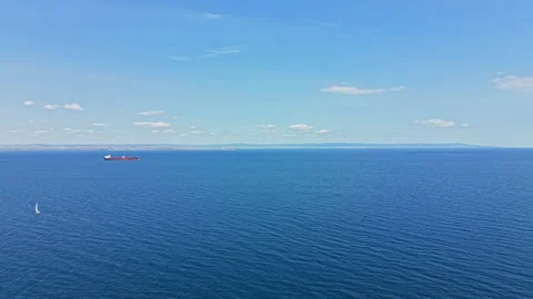 Ocean view with sailing boat and cargo ship near the coast on clear day Stock Footage 345471645
