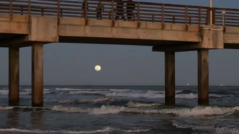 Ocean waves and moon under a pier and walkers above, in evening light. Stock Footage 236965414