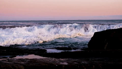 Ocean waves crash over rocky ridge into gully, peachy sunset colors, riser Stock Footage 242976394