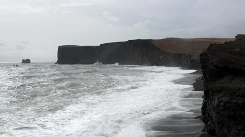 Ocean waves crashing on black sand beach with Dyrholaey cliffs under overcast Stock Footage 339303957