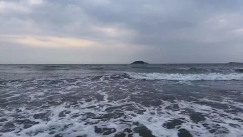 Ocean waves crashing on a sandy beach with a distant island under a cloudy sky Vidéo 334163812