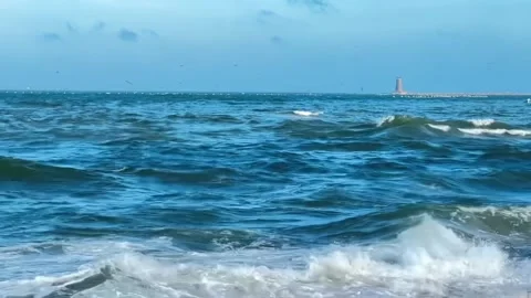 Ocean waves crashing on the shore with a distant lighthouse in the background Vidéo 334084999