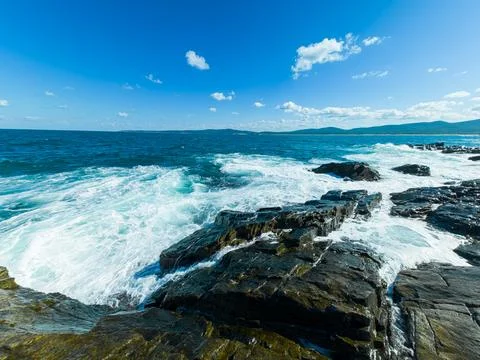 The ocean waves hit the dark rocks at the shoreline. White foam forms as the  스톡 사진