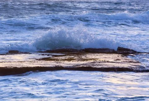 Ocean waves splash over calm rock shelf at dawn 写真素材