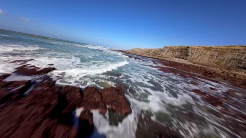 Ocean waves surge over rusty red rocks under clear skies Video stock 346077469