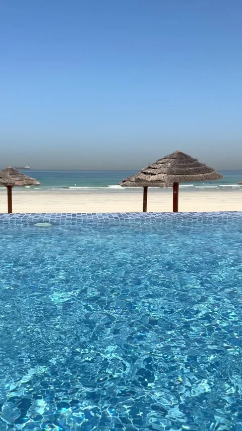 Oceanfront infinity pool beside white sand beach with sun umbrellas and lounge Stock Footage 345021544