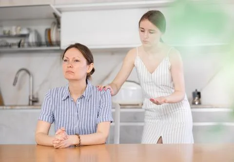 Offended mother sitting at the kitchen table, daughter standing behind Stock-Fotos