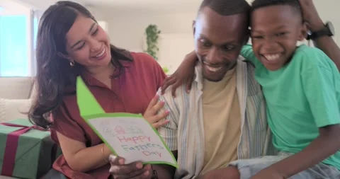 Offering mom wearing rust blouse showing Fathers Day card on sofa, dad reading, Stock Footage 340011425