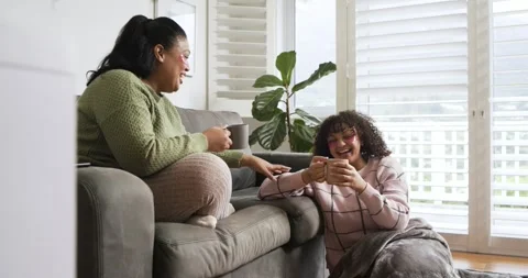 Offering mugs to share coffee, diverse female friends laughing, chatting on gray Stock Footage 339320512