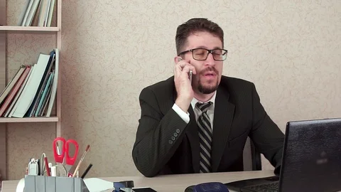Office Manager man with beard and glasses talking happily on the smartphone. 動画素材 100404002
