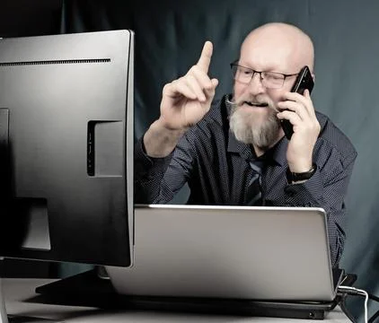 Office work - cheerful elderly man with a gray beard talking on the phone while Stock Photos