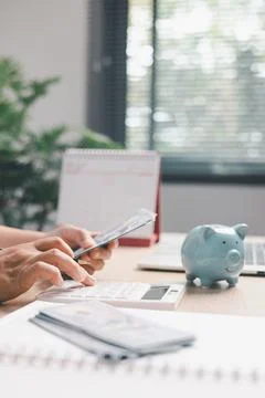 Office worker counting cash while using a calculator beside a piggy bank, i.. Stock Photos