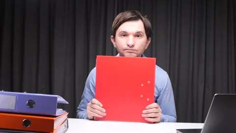 Office worker hiding behind red folder at desk with laptop and binders Stock Footage 306501921