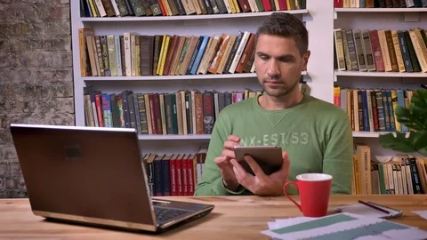 Office worker sitting in front of the laptob, scrolls down the page on a tablet Video stock 99430910