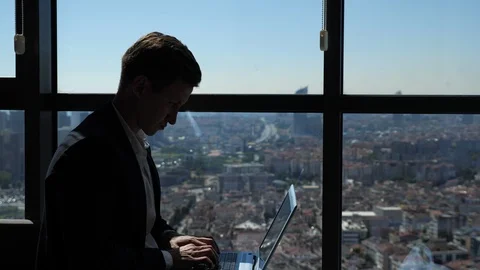 Ofiice worker in suit is typing on laptop sitting near the window with panoramic Stock Footage 111504080