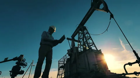 Oil industry worker walking in front of an oil derrick in sunlight. Slow motion Stock Footage 144098544