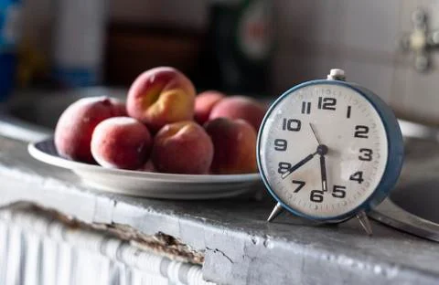 Old blue clock in front of a plate of plums marking the time for a snack Stock Photos