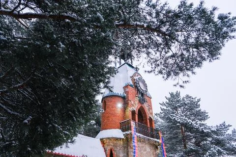 Old brick clock tower with ornate clock face covered in snow, surrounded by pine Stock Photos