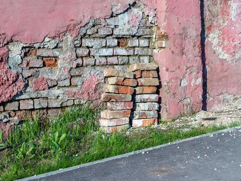 An old brick wall with peeling off plaster stands along an asphalt path, gree Stock Photos