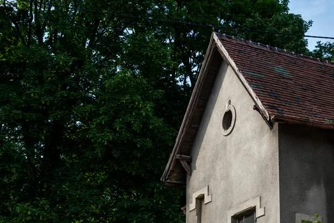 Old building with a gray facade and a triangular tiled roof next to tall trees Foto stock