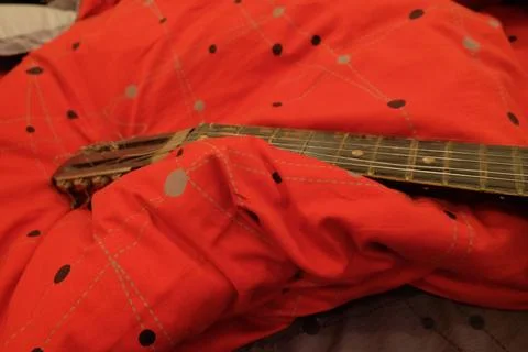 Old classical guitar on a bed in a brown apartment lying on a blanket of red Stock Photos