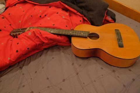 Old classical guitar on a bed in a brown apartment lying on a blanket of red Stock Photos