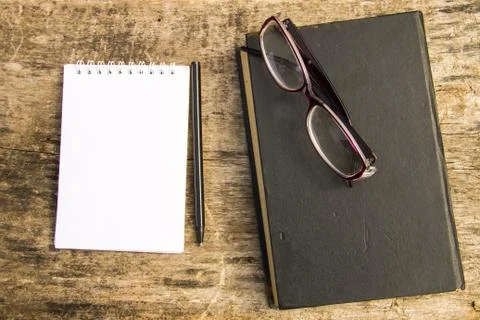 Old closed book, glasses, notepad and pen on wooden background Stock Photos