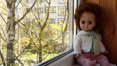 Old doll with painted eyebrows on the windowsill near the window Stock Footage 131340569