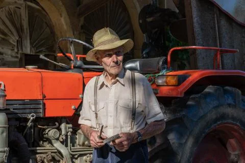 Old farmer working on red tractor under roof of farmer house in sunshine. Stock Photos