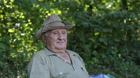 An old gray-haired man with a pensive face, in a straw hat sits on a chair in th Stock Footage 120166123