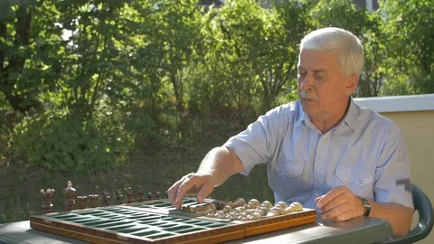 Old gray-haired man prepares the chess game outside in the garden at the table Stock Footage 118529645