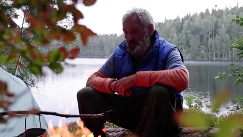 An old grey-haired man setting a fire near the river. Stock Footage 235886327
