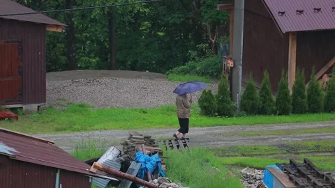 An old lady walking through the rain in a mountain village holding an umbre.. Vídeos de archivo 329063876
