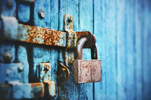 An old large lock hangs on a rust-covered blue abandoned wooden gate. Closed. Stock Photos