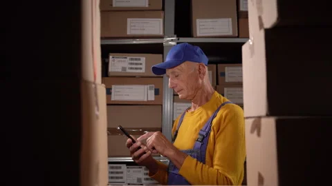 Old male warehouse worker using smartphone while working with delivery and Stock Footage 249597175
