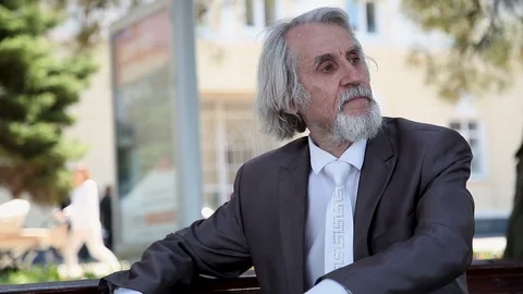 Old man with beard in suit, tie sitting on Park bench looking around Vidéo 96411675
