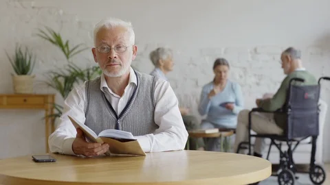Old man with book looking at camera while friends playing cards Stock Footage 117575033