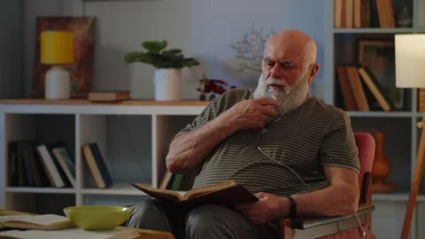 Old man in breathing mask stay in chair and read book. Old man is using Stock Footage 247466341
