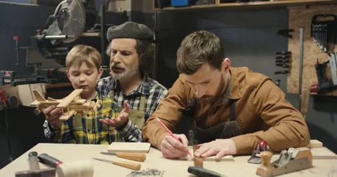 Old man carpenter sitting at desk in workshop with handsome son and cute little Stock Footage 130363772