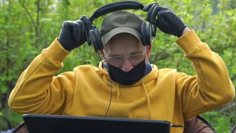 Old man in face mask and gloves puts on headphones at laptop Stock Footage 130888293