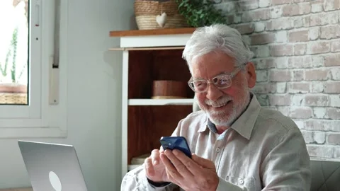 Old man in glasses sit in kitchen near laptop holding smartphone device cha.. Stock Footage 236928235