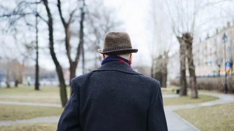 Old man in hat is walking in city park, back view. He limps on his leg, lame man Stock Footage