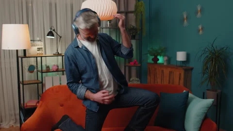 Old man in headphones dancing on sofa at home listening favorite music, playing Stock Footage 230116780
